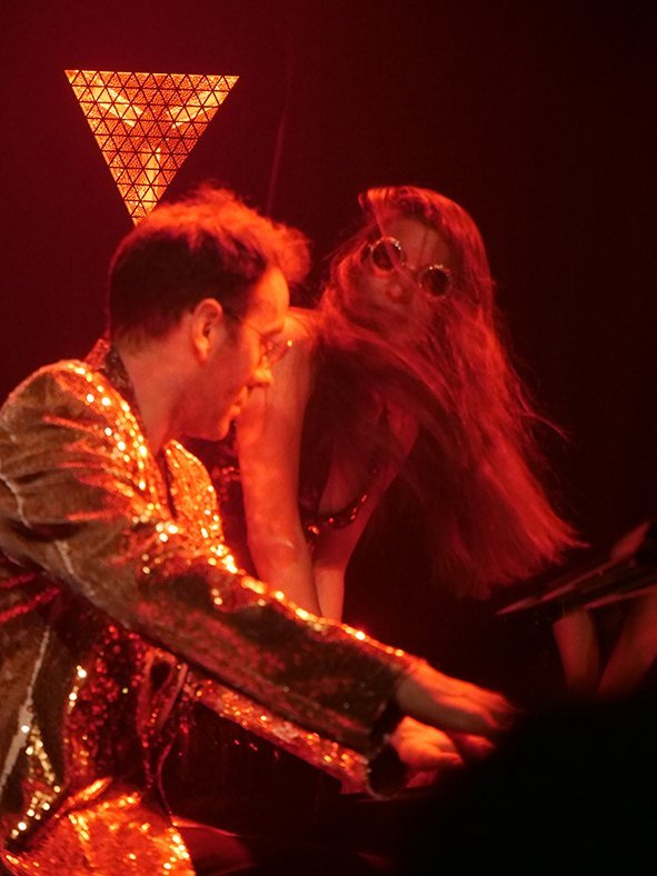 Dexter en veste à paillettes et Louise en lunettes rondes sur scène, sous une lumière rouge
